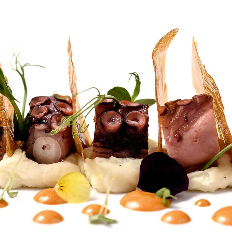Plato gourmet con pulpo, montado sobre puré, con crujientes y salsas decorativas en un arreglo elegante.