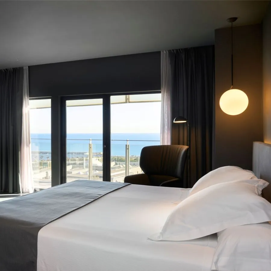 Habitación luminosa con cama doble, lámpara esférica y ventana con vista al mar.