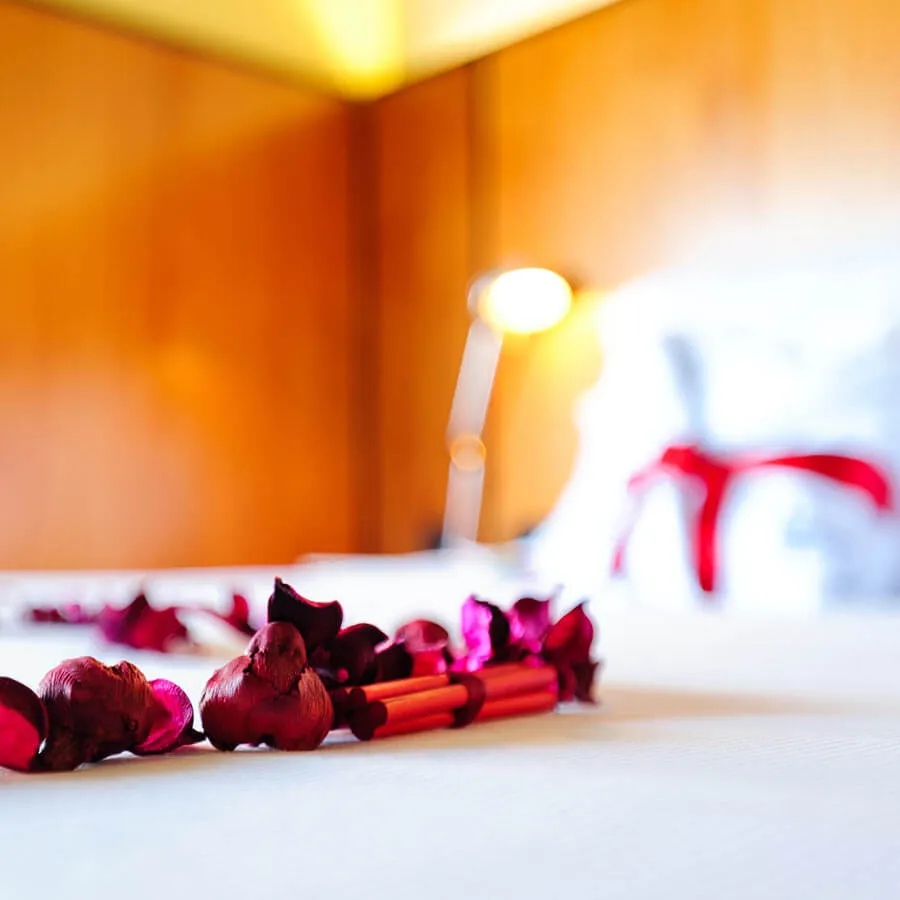 Pétalos rojos sobre una cama blanca y lámpara de lectura en habitación de madera. Ambiente romántico y cálido.