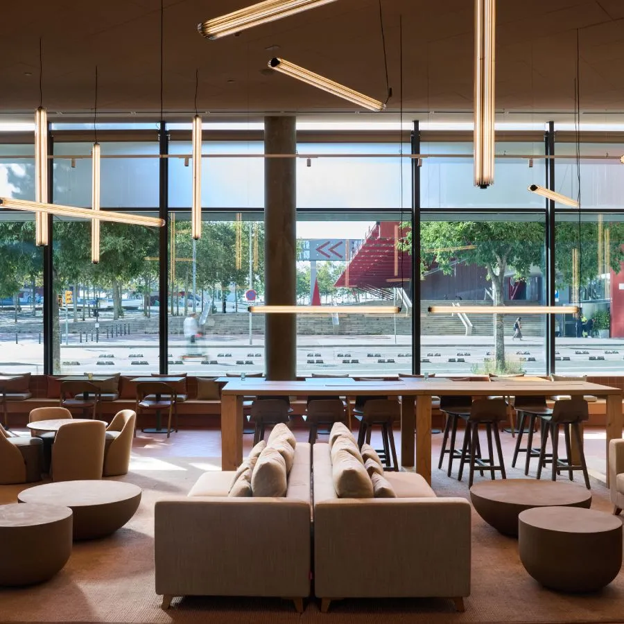 Sala moderna con sofás beige, mesas y lámparas colgantes cerca de grandes ventanas que dan a una calle arbolada.