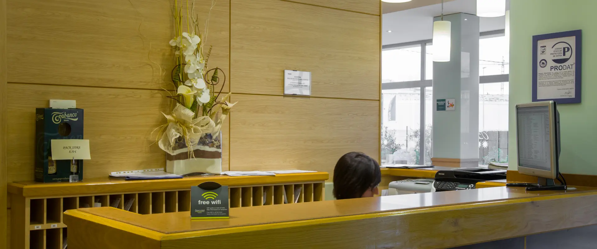 Recepción con mostrador de madera, computadora, y jarrón decorativo. Letrero de wifi gratis.