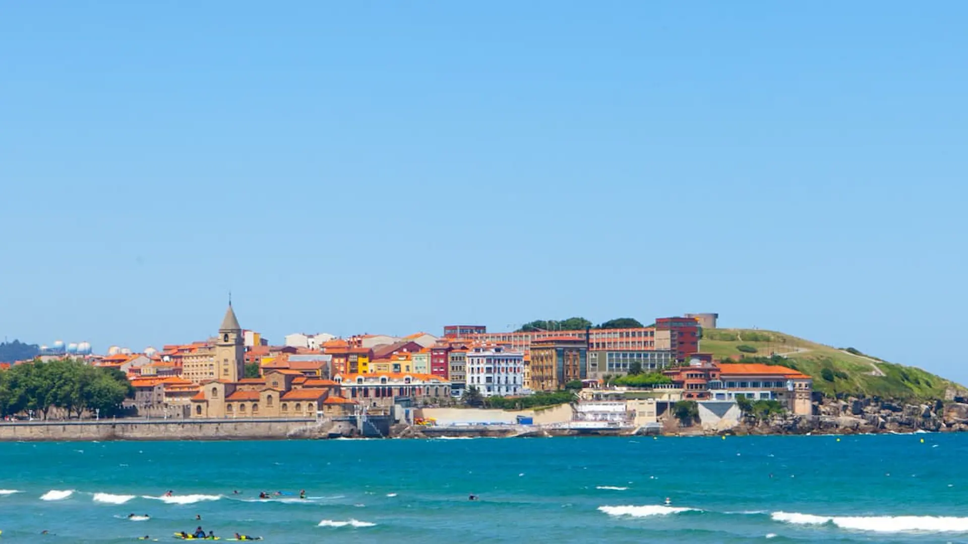 Vista de una ciudad costera con edificios coloridos y una iglesia, frente al mar azul.