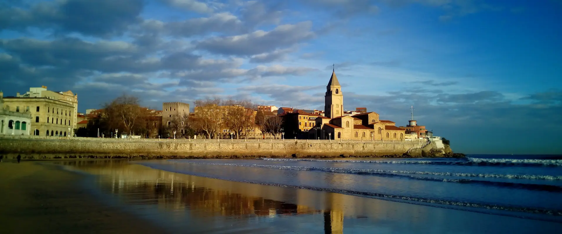 Vista de una iglesia y edificios históricos junto al mar bajo un cielo parcialmente nublado.