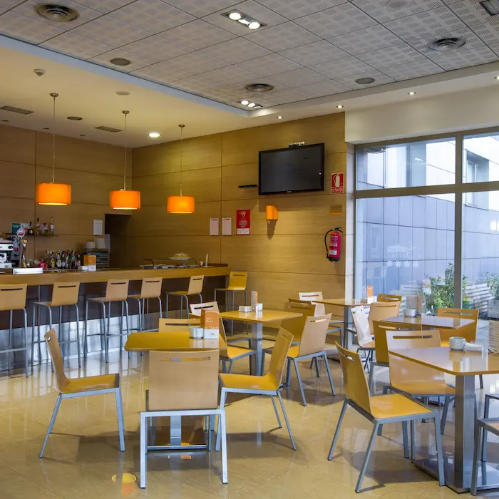 Cafetería moderna con mesas y sillas de madera, barra y lámparas naranjas colgantes.