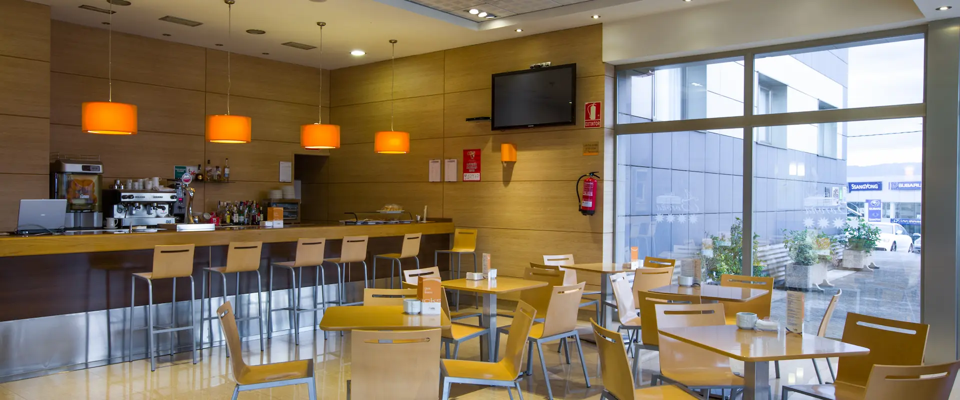 Cafetería moderna con sillas de madera, barra de bar y lámparas naranjas colgantes.