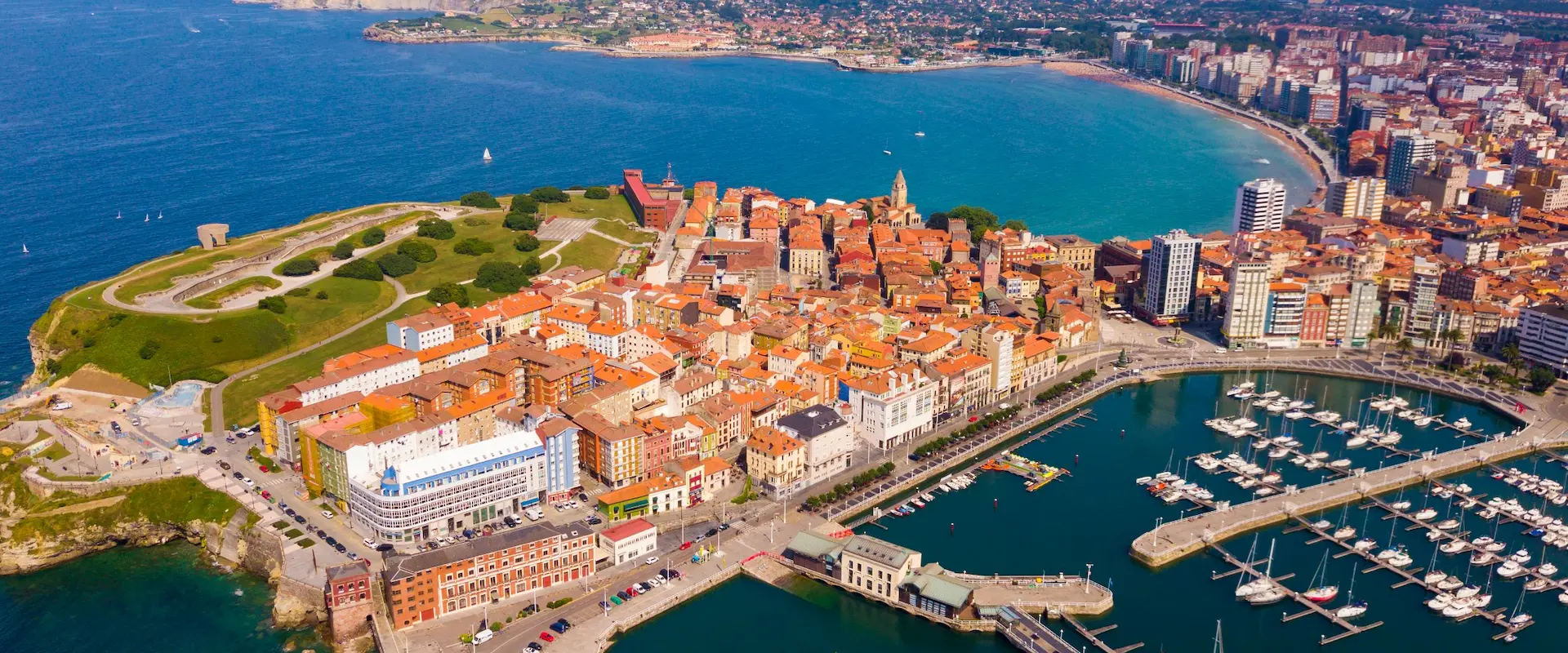 Vista aérea de una ciudad costera con puerto, edificios de tejados rojos y mar azul en el fondo.