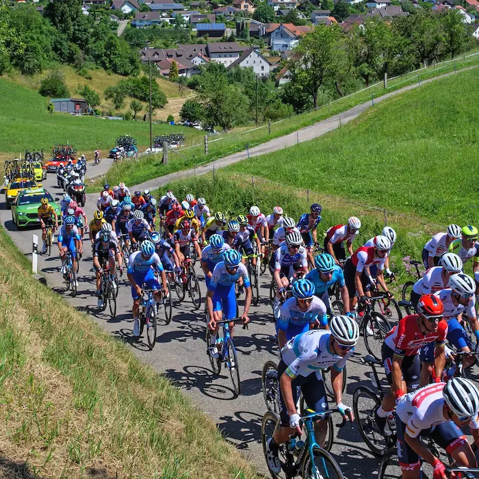 Ciclismo en grupo subiendo una colina, rodeados de coches de equipo y un paisaje verde.