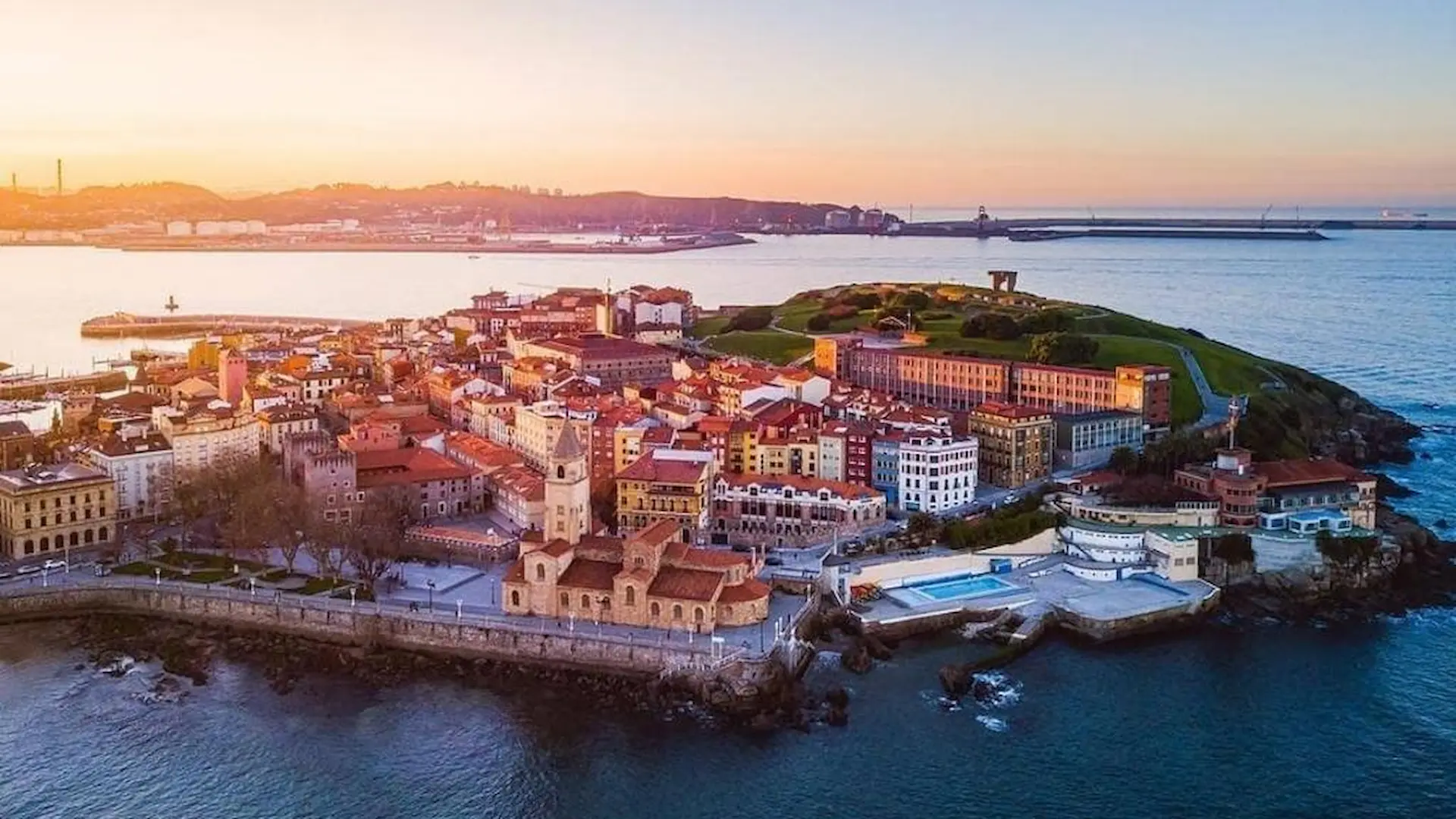 Vista aérea de un paisaje costero al atardecer, con edificios y una iglesia cerca del mar.