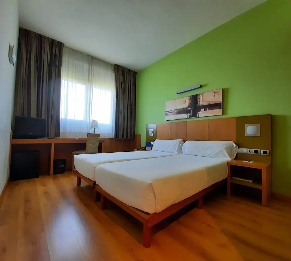 Habitación de hotel con dos camas, pared verde y ventana con cortinas.