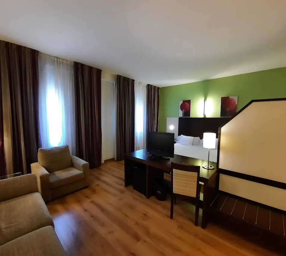 Habitación de hotel con cama, sofá, escritorio, televisor y decoración moderna en tonos cálidos.