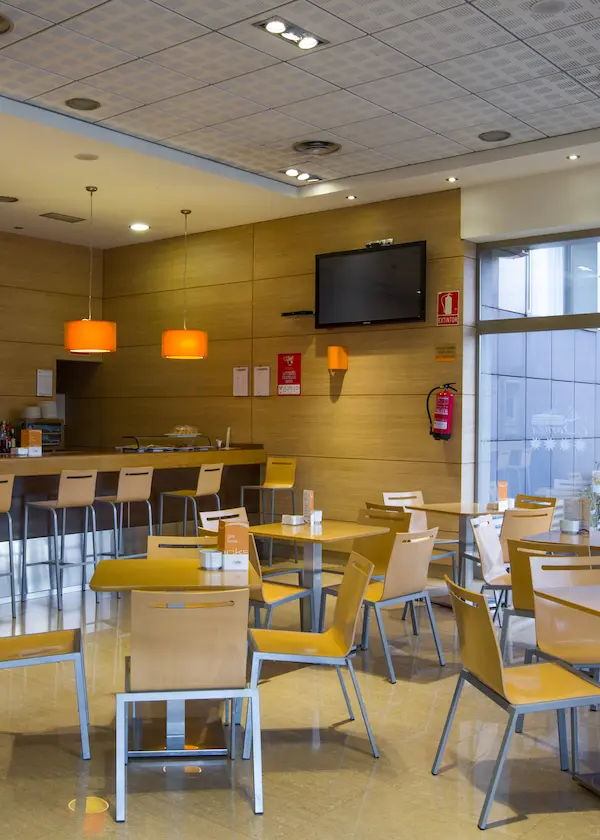 Cafetería moderna con sillas y mesas de madera clara, lámparas naranjas y televisor en la pared.