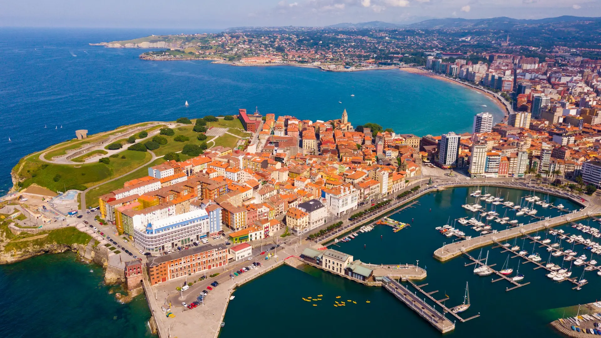 Vista aérea de una ciudad costera con puerto, edificios y un paisaje verde cerca del mar.