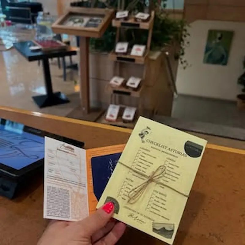 Persona sosteniendo papeles en un mostrador, fondo con folletos en un estante de madera.