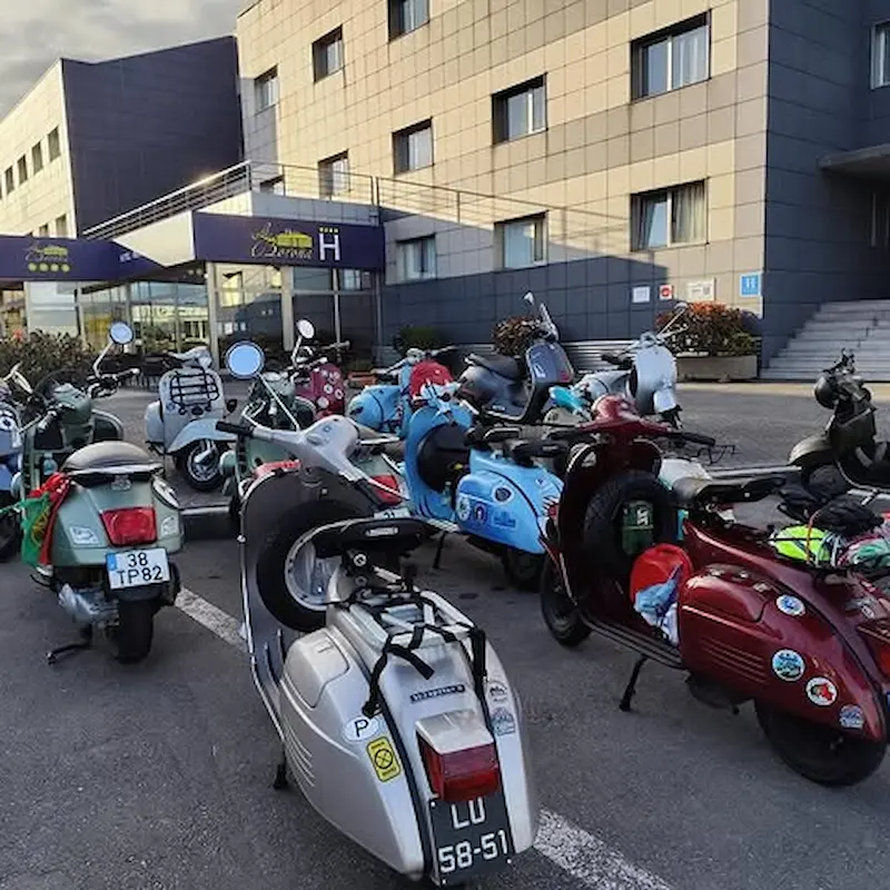 Una fila de scooters vintage estacionadas frente a un hotel moderno con fachada gris.