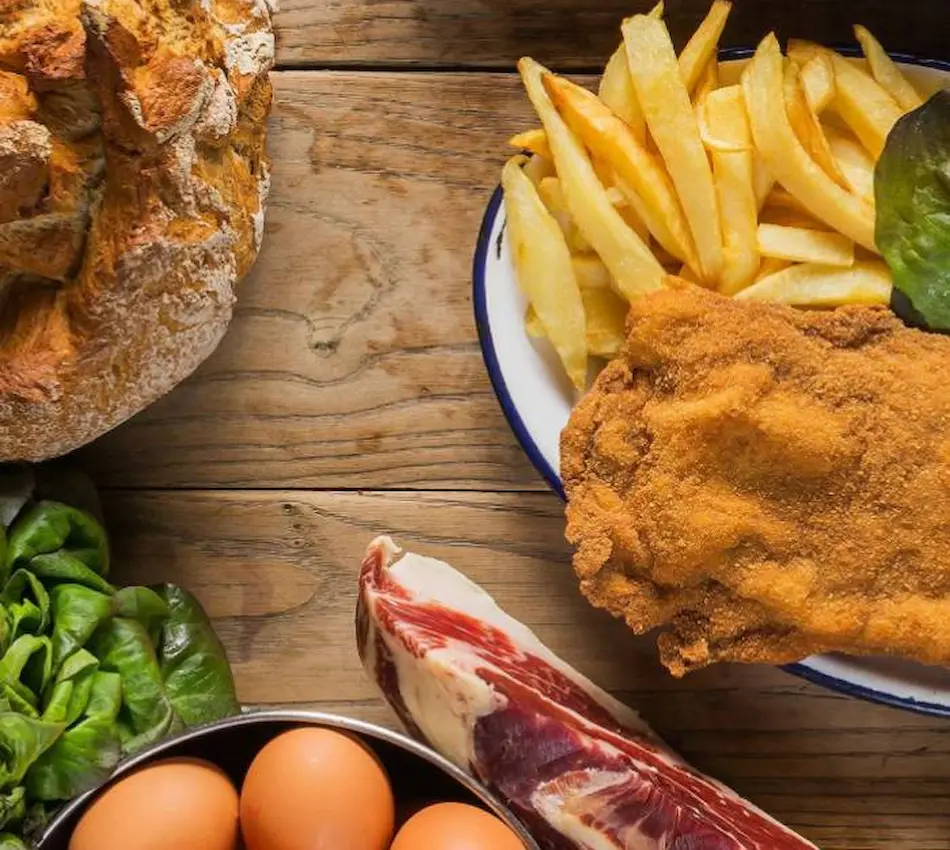 Plato de milanesa con papas fritas, pan, huevos, lechuga y jamón sobre mesa de madera.