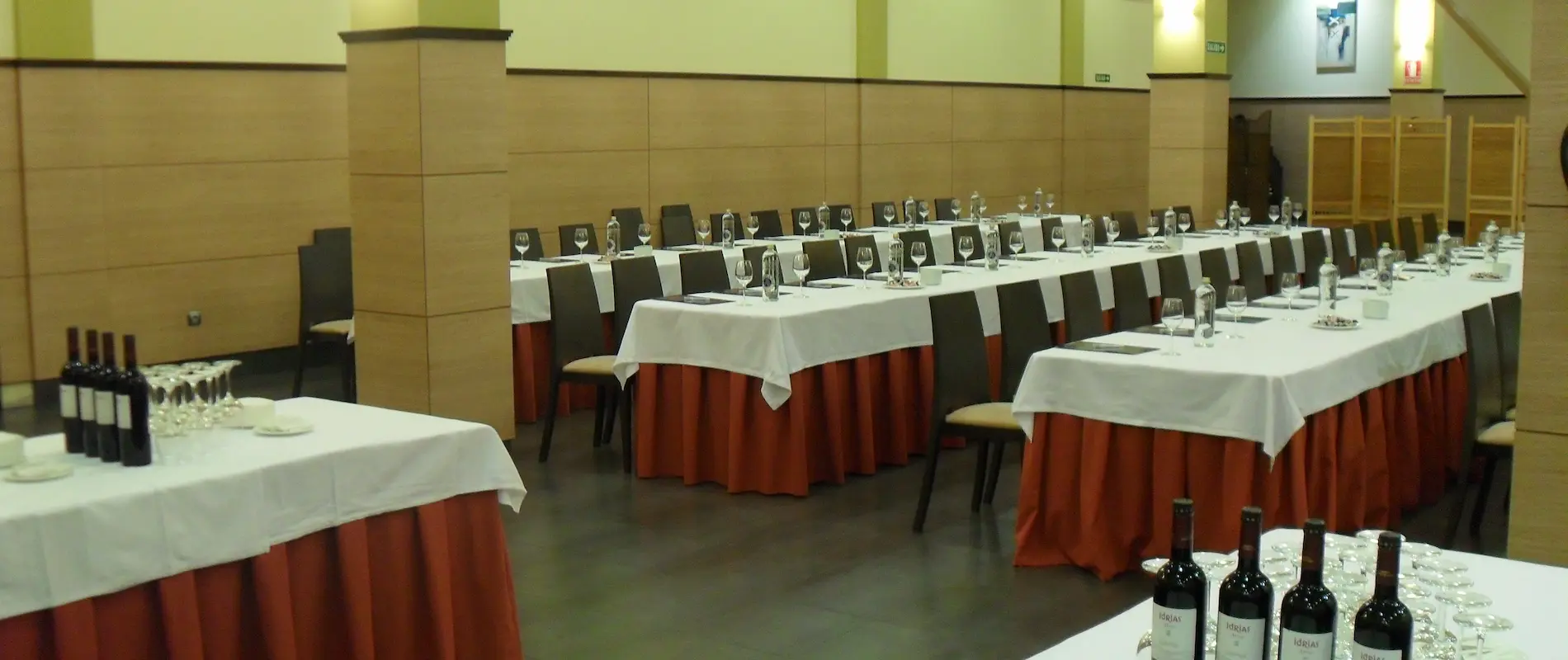 Sala de conferencias preparada con mesas, copas y botellas de vino bajo luz cálida.