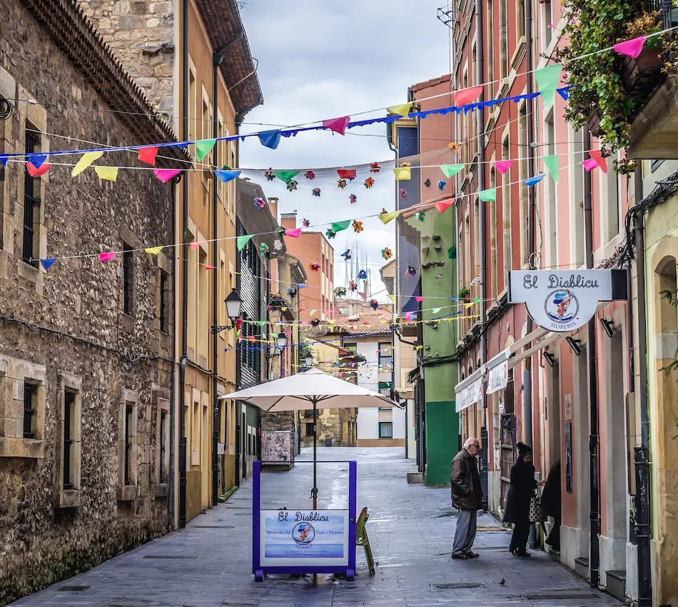 Calle estrecha con decoraciones coloridas y tiendas a los lados bajo un cielo nublado.