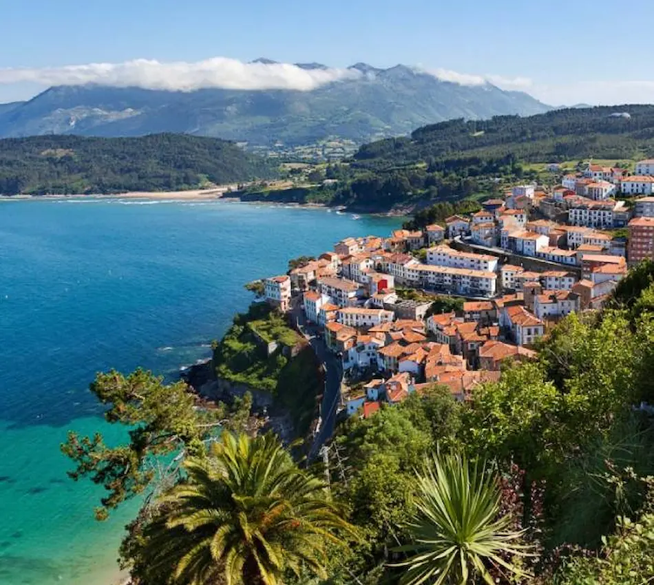 Costa con pueblo y mar azul, montañas al fondo, cielo con nubes y vegetación verde.