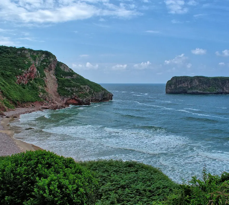 Costa rocosa con mar azul y cielo despejado, rodeada de vegetación verde y acantilados.