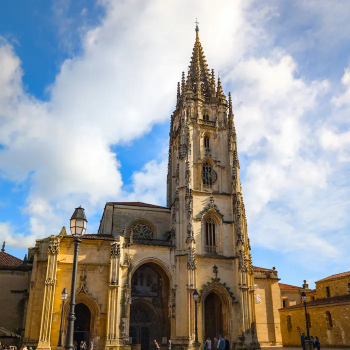 Torre de una catedral gótica contra un cielo azul y nubes, con detalles arquitectónicos ornamentados.