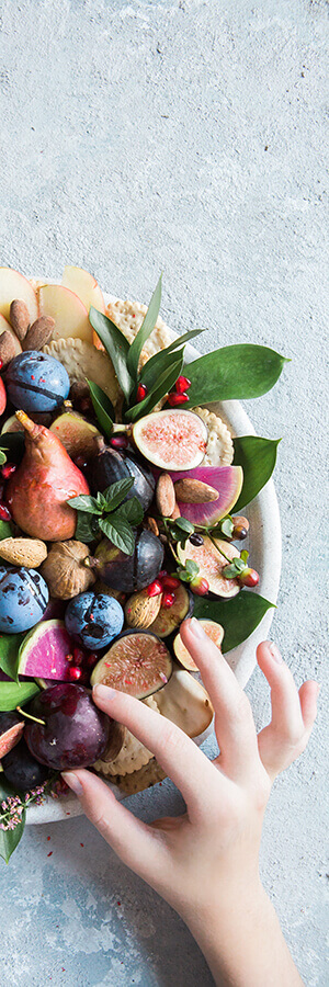 Mano cerca de una bandeja con frutas variadas, nueces y hojas verdes sobre fondo gris.
