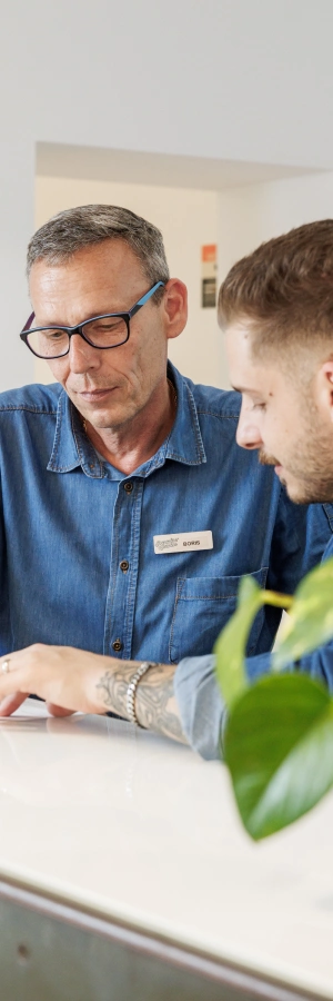 Dos hombres conversando frente a un escritorio blanco con planta verde.