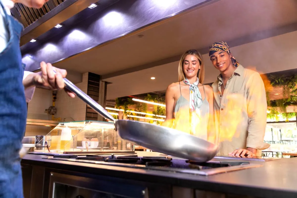 Persona cocina frente a dos personas sonrientes; sartén con llamas en una cocina abierta.