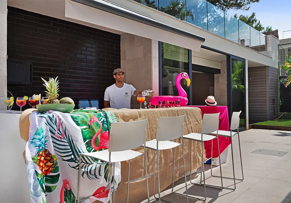 Bar al aire libre con decoración tropical, cócteles y un flamenco inflable. Persona de pie detrás.
