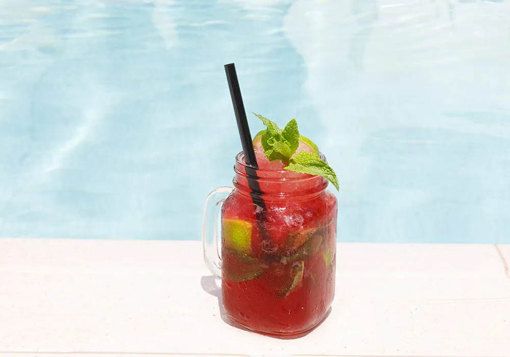 Jarra de vidrio con bebida roja, hielo, limón y menta, junto a una piscina.