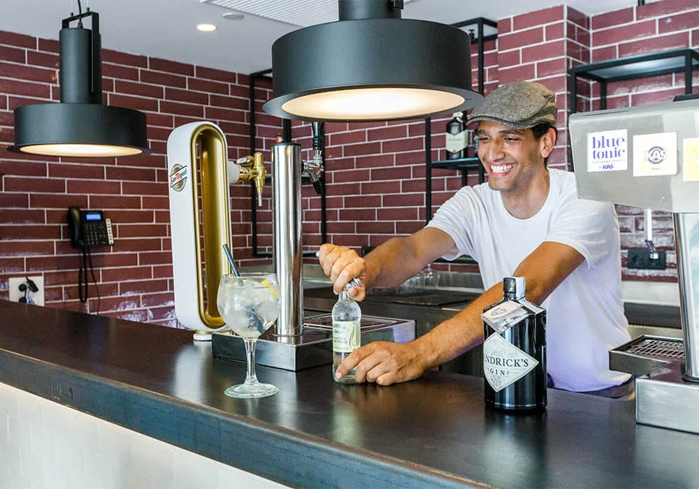 Persona sonriendo detrás de una barra, sirviendo bebida. Hay botellas de ginebra y tónica.