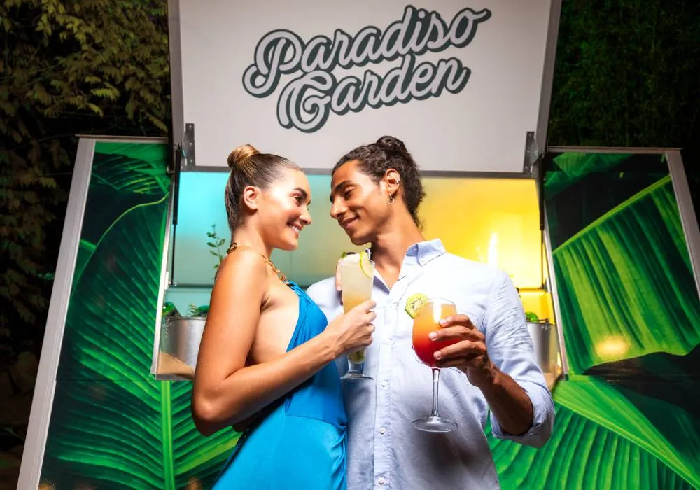 Pareja sonríe con bebidas en mano, bajo letrero de "Paradiso Garden" con fondo de hojas verdes.