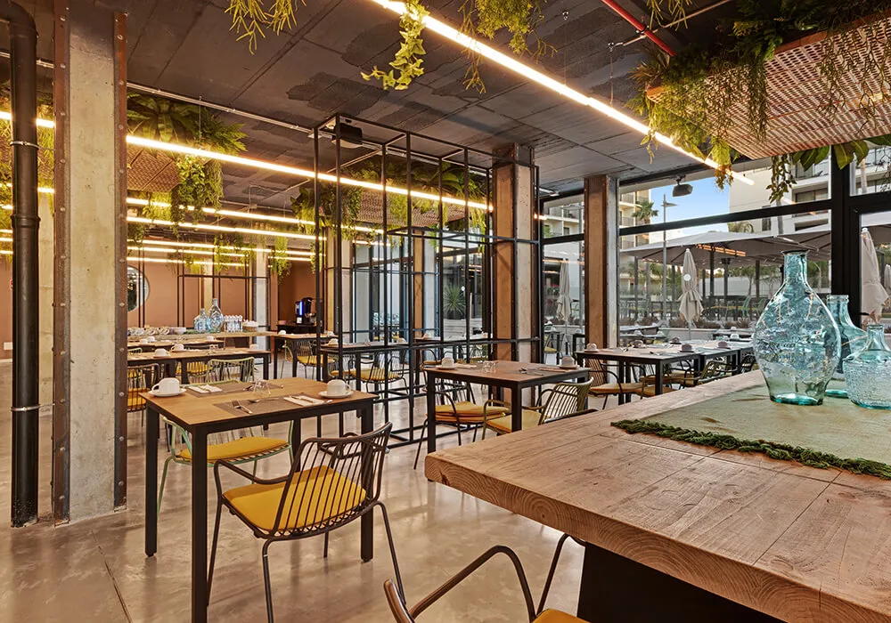 Restaurante moderno con mesas de madera, sillas amarillas y plantas colgantes.