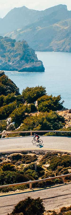 Ciclista en carretera costera con montañas y el mar de fondo.