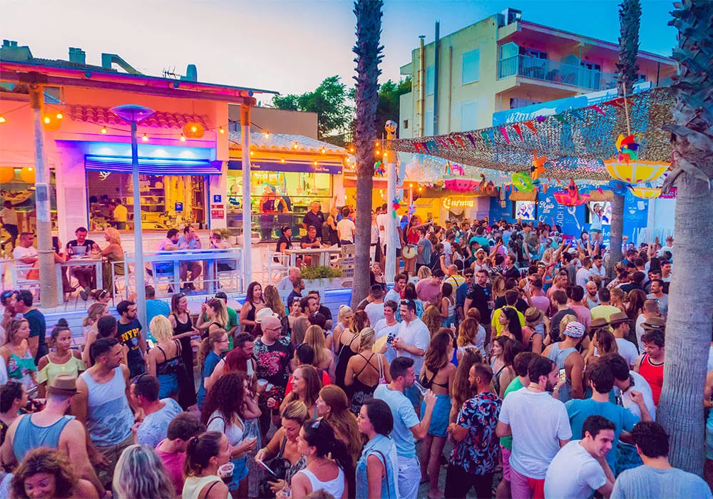 Multitud disfrutando de una fiesta al aire libre con luces, música y decoración colorida.
