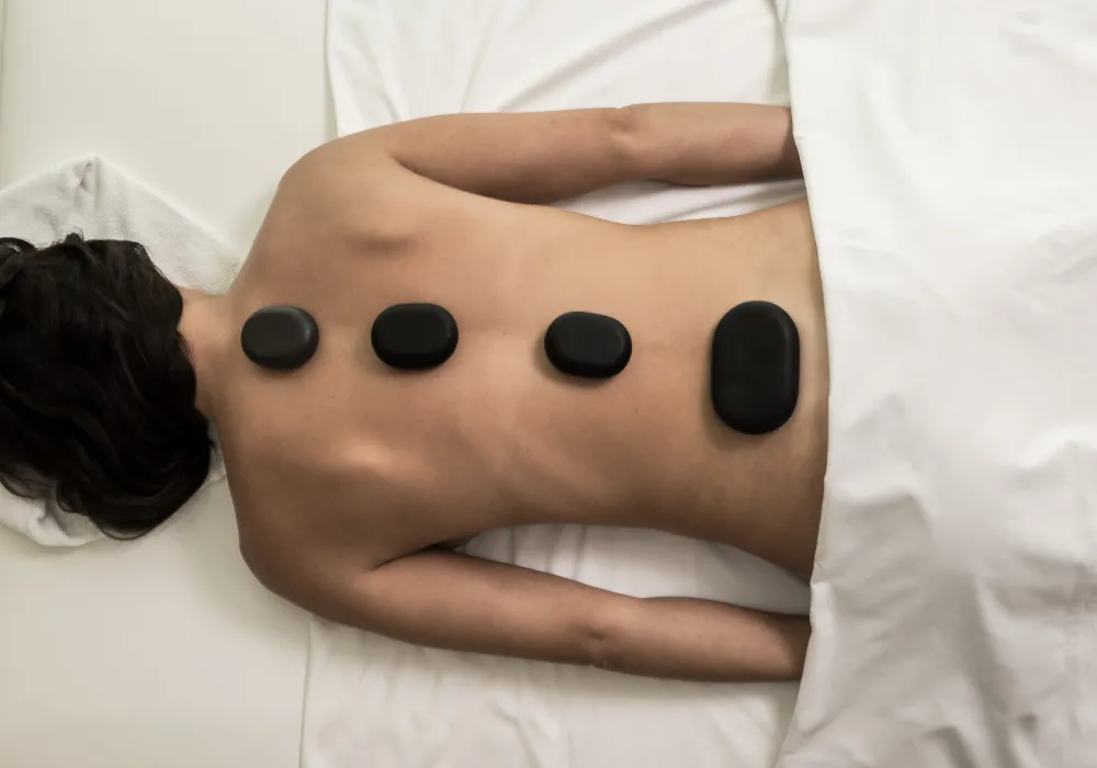 Persona recibiendo terapia de piedras calientes sobre una cama blanca.