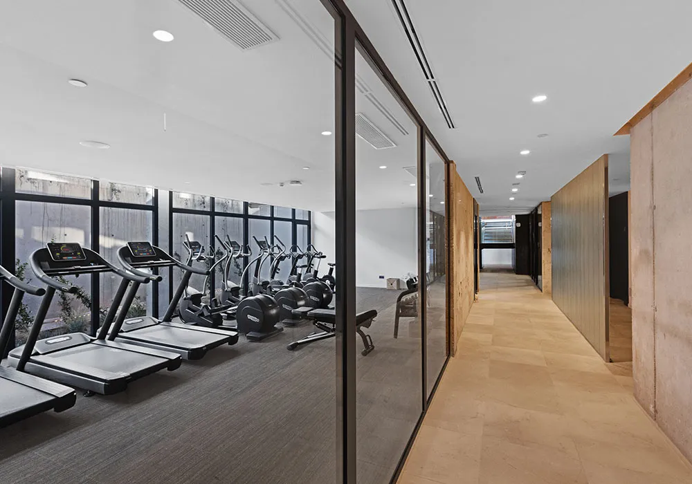 Gimnasio moderno con cintas de correr y bicicletas junto a un pasillo con paredes de vidrio.