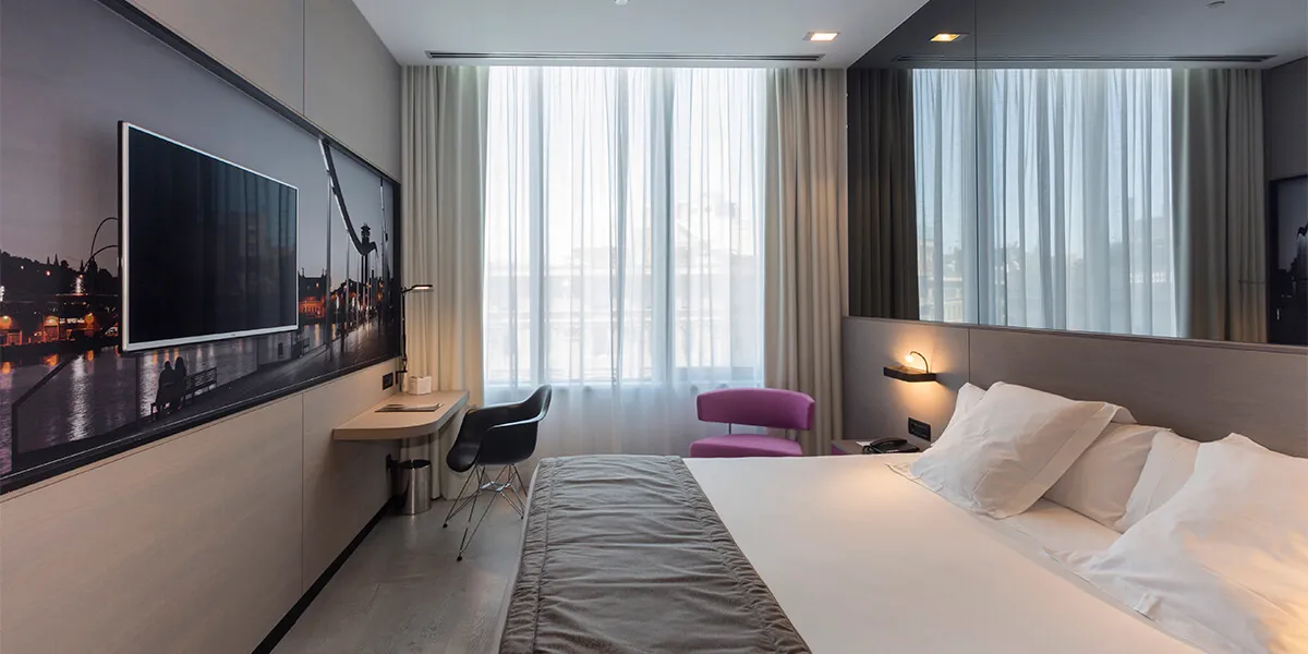 Habitación moderna con cama doble, escritorio, TV y ventana grande con cortinas blancas.