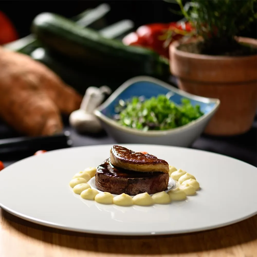 Plato gourmet con carne y puré, fondo de vegetales y plantas decorativas.