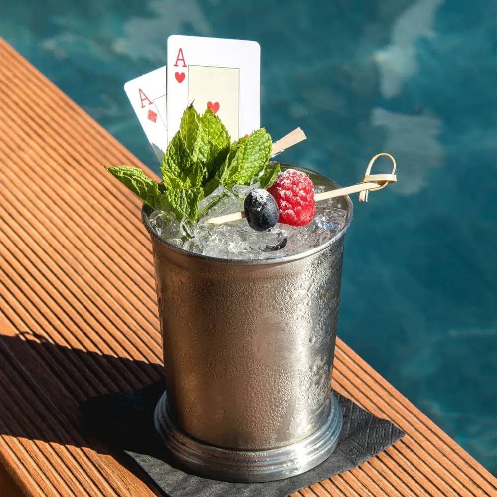 Cóctel en vaso de metal con menta, frambuesa, arándano, hielo y dos ases junto a una piscina.