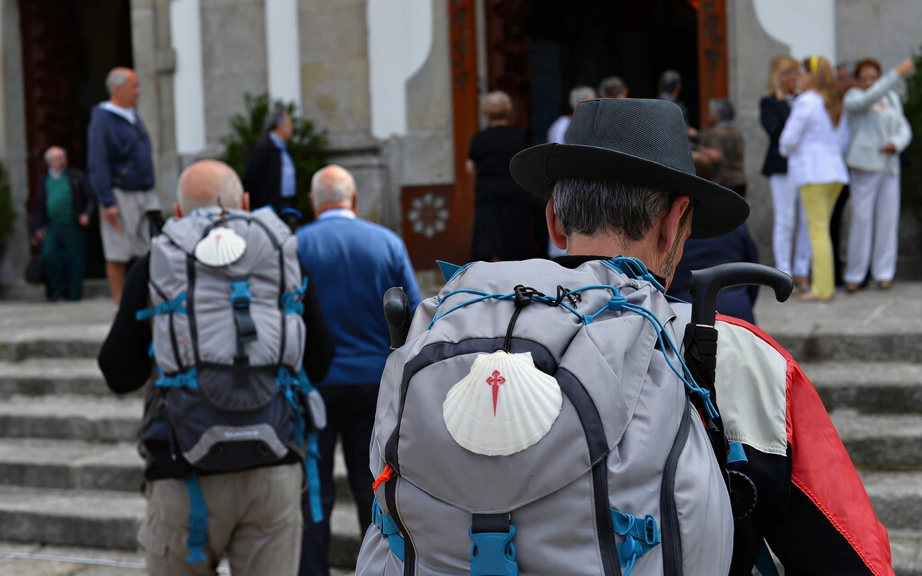 Pilgrims with backpacks and scallop shells, heading toward a building entrance.