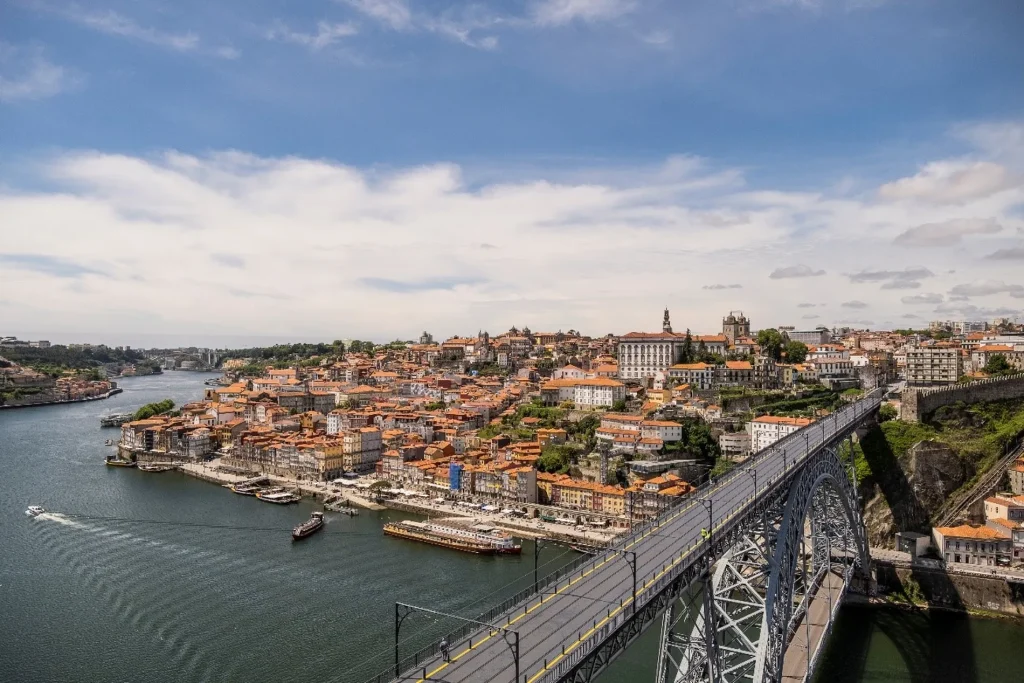 Cityscape of Porto, Portugal with Douro River and Dom Luís I Bridge under a partly cloudy sky.