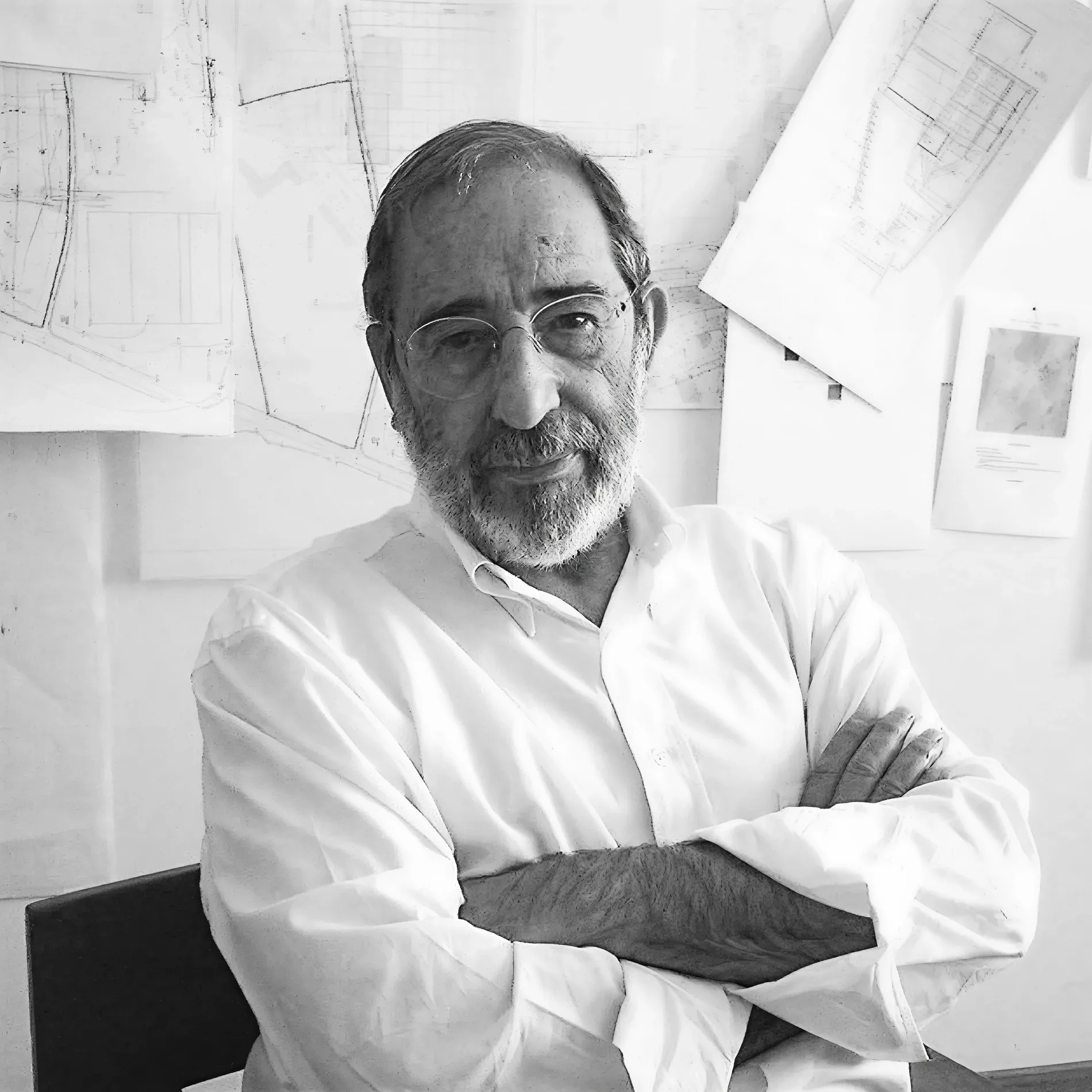 Bearded man in glasses with arms crossed, standing in front of architectural drawings.