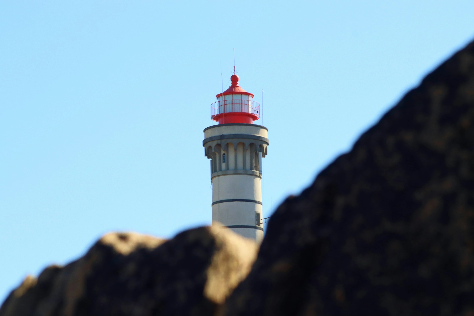 Lighthouse with red top viewed between dark rocks against a clear blue sky.