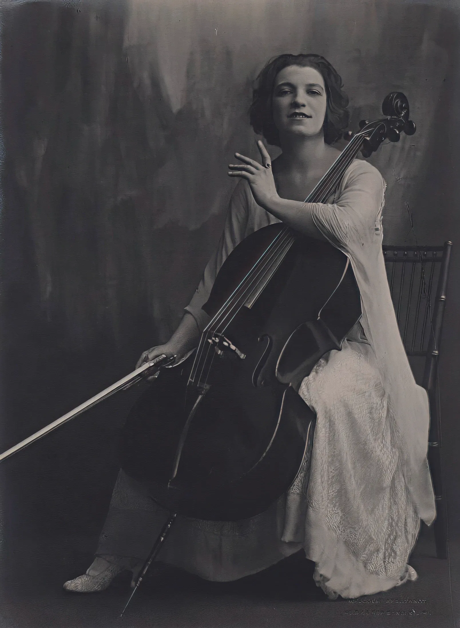 A woman seated with a cello, poised to play, in a vintage black and white photograph.
