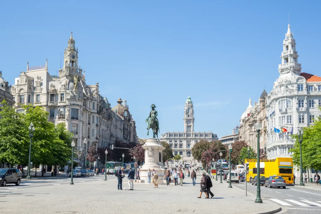 City square with historic buildings, statue, people, cars, and a clear blue sky.