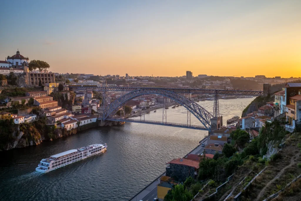 River at sunset with a distant bridge, boat, and hillside buildings.