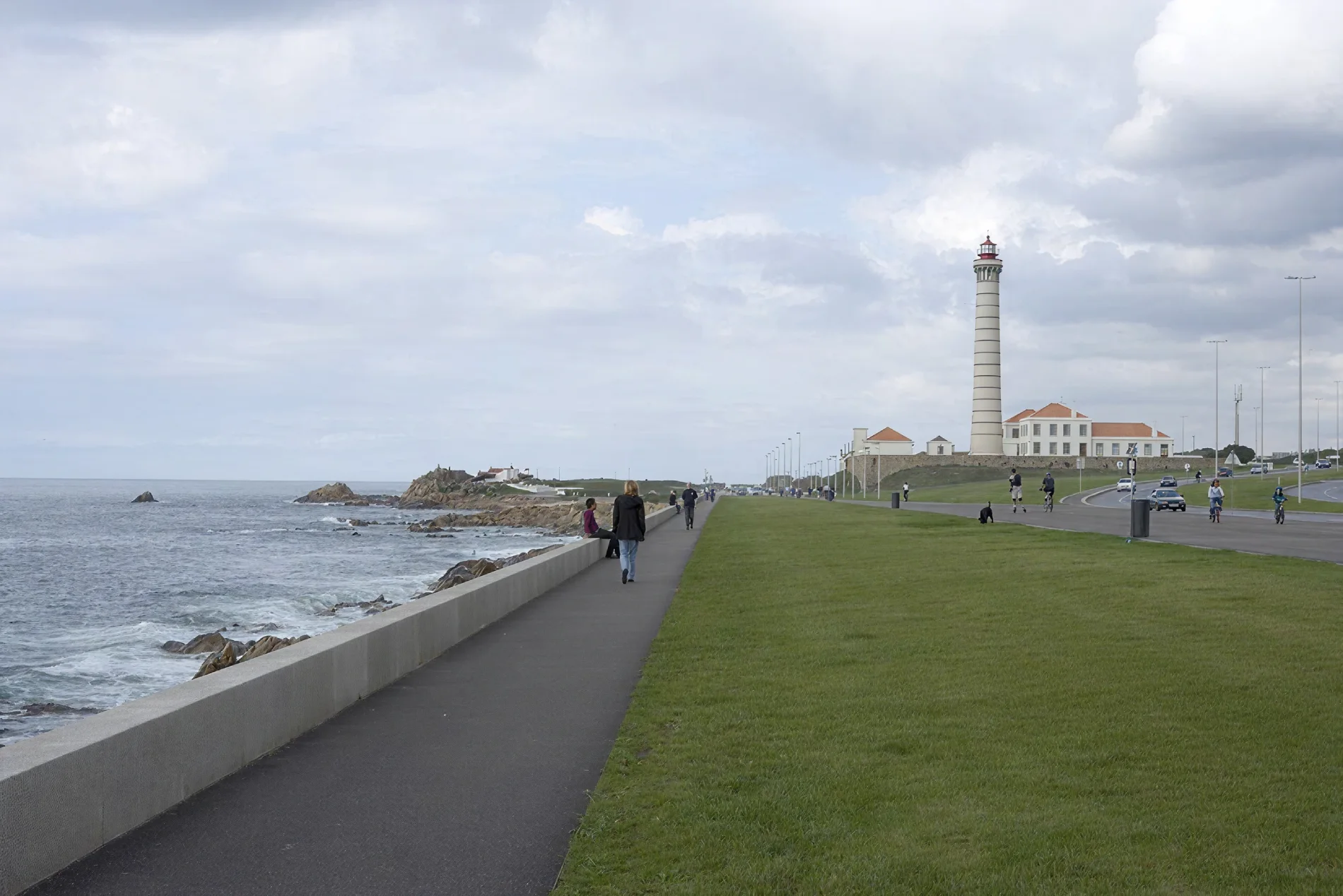 Coastal path with people walking, near a lighthouse and grassy area. Cloudy sky above.