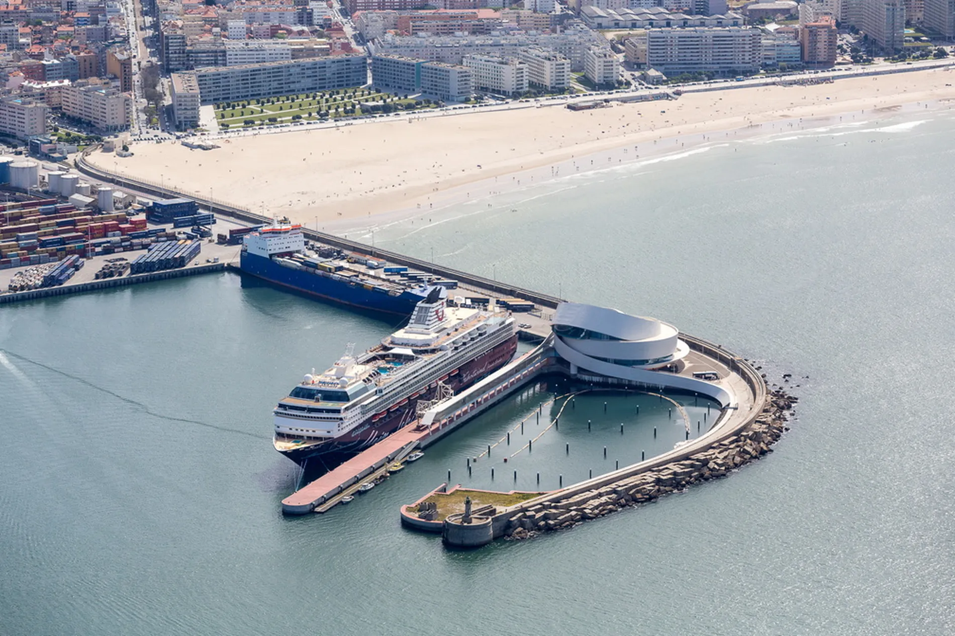 Aerial view of a cruise ship docked at a modern port near a beach and cityscape.