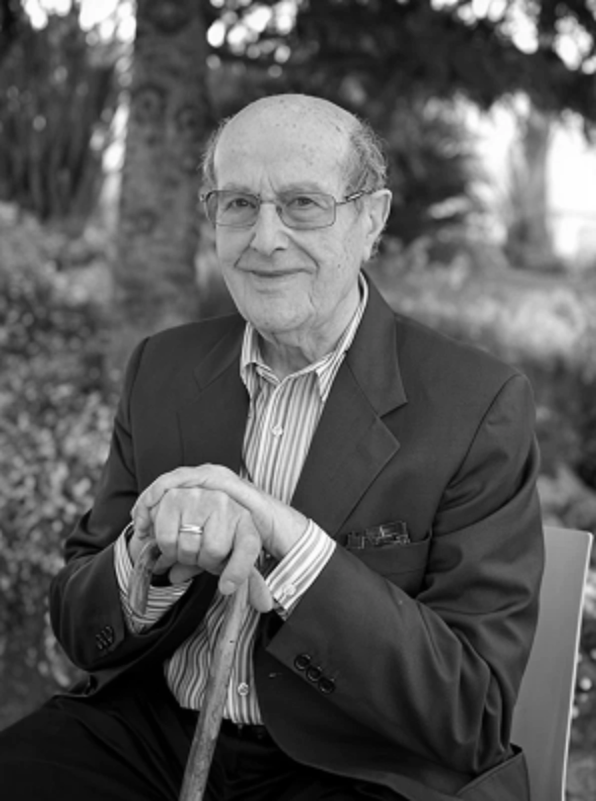 Elderly man in a suit, sitting outdoors with a cane, smiling.