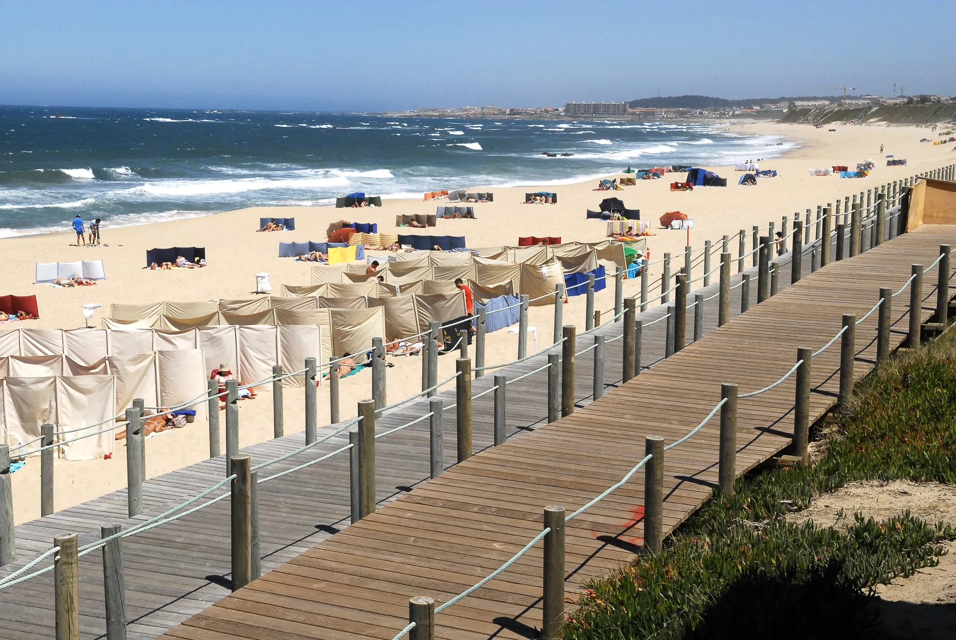 Sunny beach with people, privacy screens, and a wooden boardwalk by the ocean.